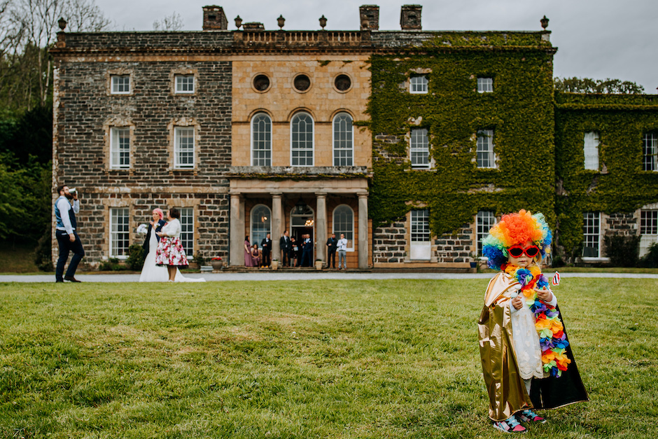 Eye-catching portrait of little girl in rainbow-colored wig and gold cape in front of mansion.