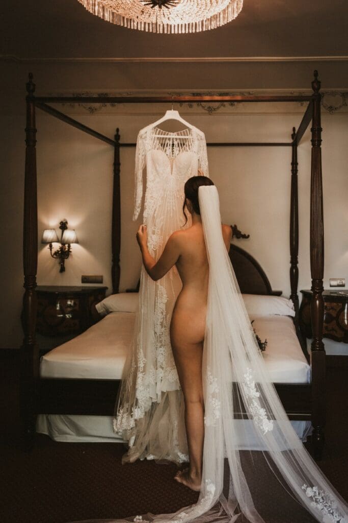 Bride with veil and no clothes on looks at her wedding dress hanging on bed rail.